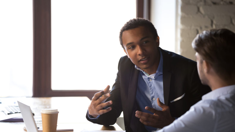 Focused African American male businessman talk with colleagues, explain business project sitting at table, diverse employees negotiate in office, discussing business issues or contract terms