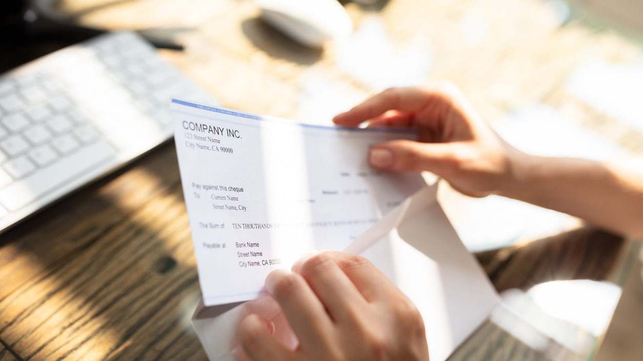 Close-up Of A Businessperson's Hand Opening Envelope With Paycheck Over Wooden Desk