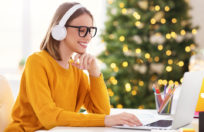 Happy pretty young woman freelancer in headphones smiling, working remote at laptop at home before Christmas