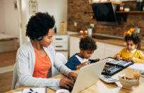 Happy African American single mother working on a computer while the kids are drawing beside her at home.