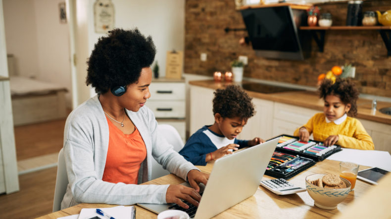 Happy African American single mother working on a computer while the kids are drawing beside her at home.
